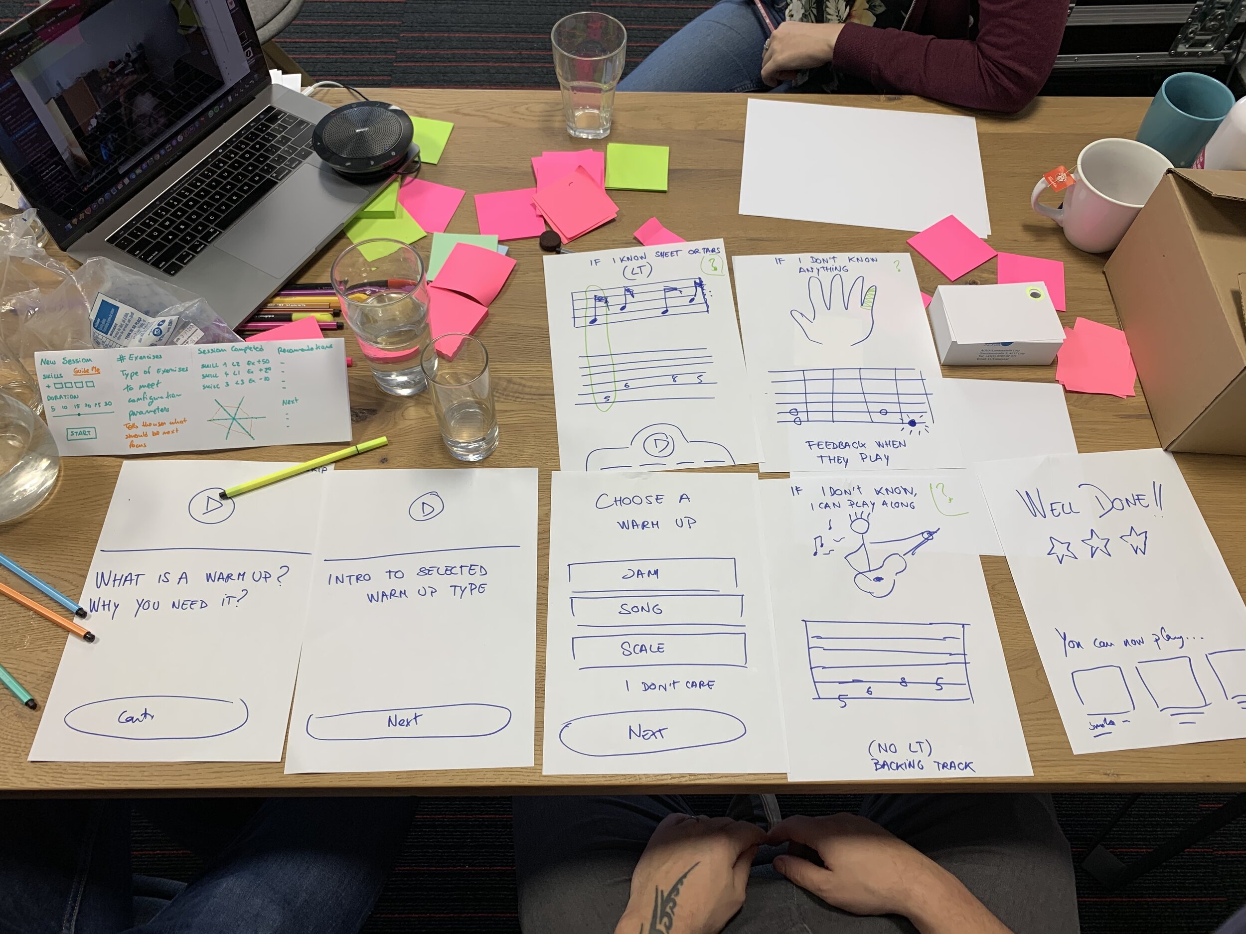 A Fretello workshop table covered in hand-drawn paper prototypes: warm-up screens, fretboard sketches, 'WELL DONE' celebration screens, and pink and green sticky notes scattered across a wooden table.